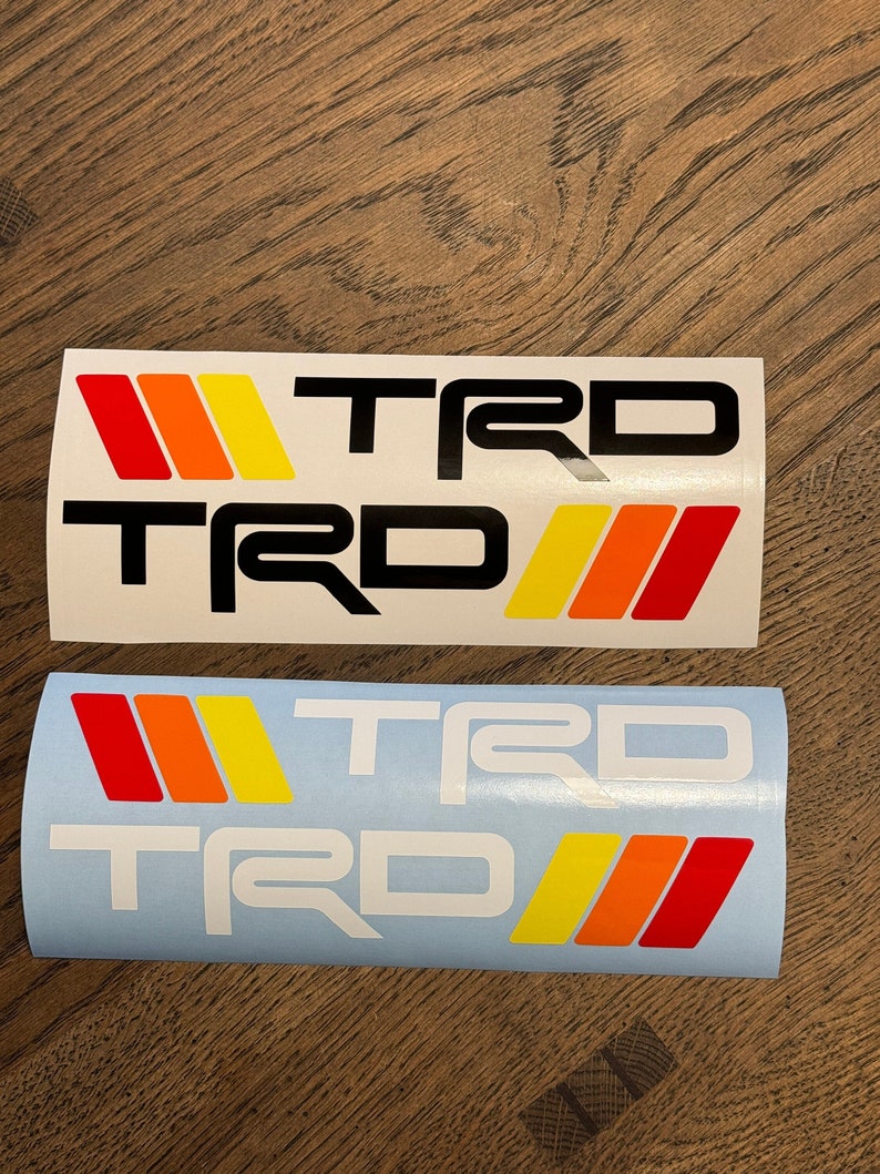 May include: Two white decals with the word "TRD" in black letters and a red, orange, and yellow stripe design. The top decal is on a white background and the bottom decal is on a light blue background.