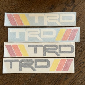 May include: Four white stickers with the word "TRD" in black and white lettering. The stickers have red, orange, and yellow stripes.