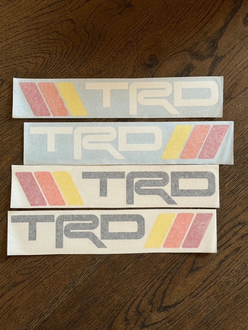 May include: Four white stickers with the word "TRD" in black and white lettering. The stickers have red, orange, and yellow stripes.