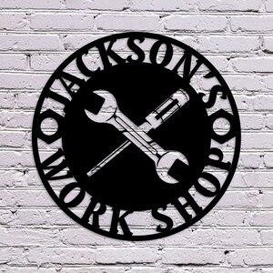 May include: Black metal sign with the words "Jackson's Workshop" in a circular design. The sign features a wrench and screwdriver crossed in the centre, set against a black background. The sign is mounted on a white brick wall.
