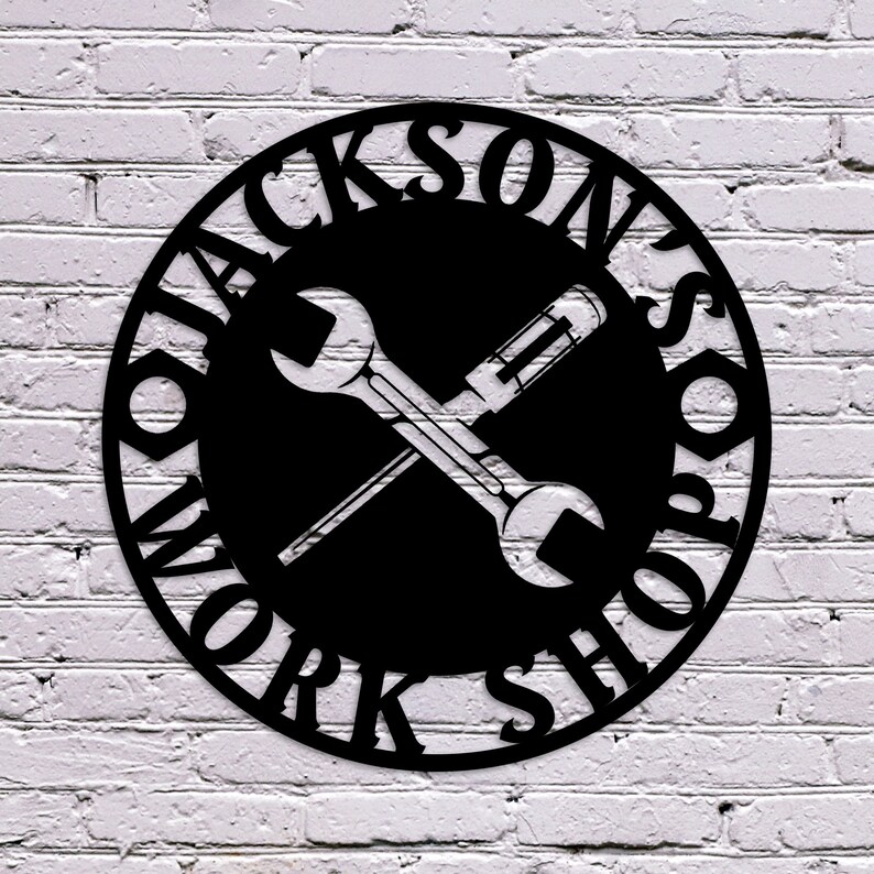 May include: Black metal sign with the words "Jackson's Workshop" in a circular design. The sign features a wrench and screwdriver crossed in the centre, set against a black background. The sign is mounted on a white brick wall.