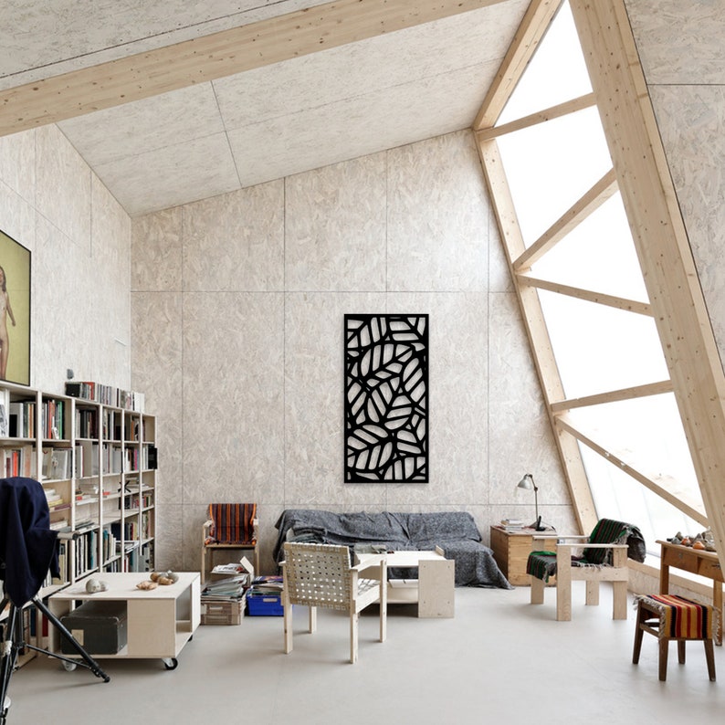 May include: A modern living room with a high, angled ceiling and large window. The room features a black, leaf-patterned wall art, a gray sofa, and light-colored wooden furniture. Bookshelves and a small table with a lamp are also visible.