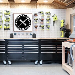 May include: A workshop interior with a framed black metal sign reading "JACKSON'S WORK SHOP" with crossed wrenches and a screwdriver. Black tool chests are below. Power tools are mounted on a pegboard wall, with a band saw visible.