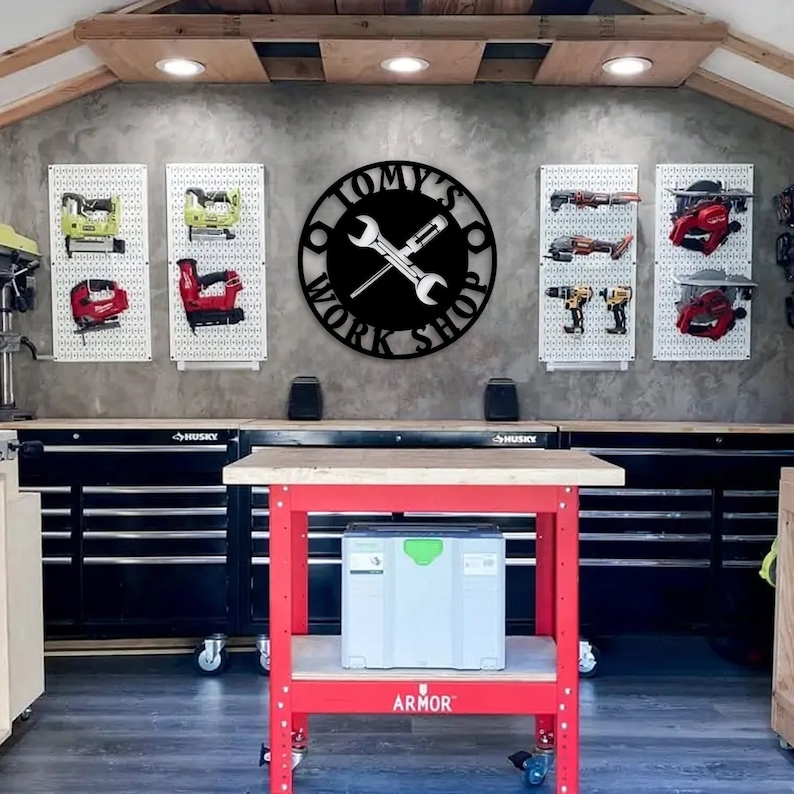 May include: A workshop interior with a red workbench and a sign that reads "TOMY'S WORK SHOP". The sign features crossed wrenches and a screwdriver. Pegboards on the wall display various tools. The workbench is red and has a wooden top.