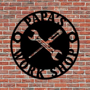 May include: Black metal sign with the words "PAPA'S WORK SHOP" in a circular design. The sign features crossed wrenches in the centre and is mounted on a red brick wall.