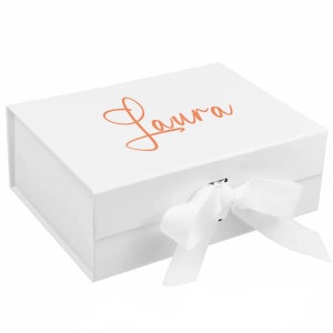 May include: White gift box with a white ribbon and the name "Laura" written in orange on the top.
