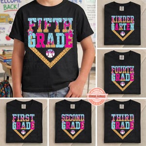 May include: A black short-sleeve t-shirt featuring a graphic of a baseball home plate with the text FIFTH GRADE in light blue and pink. The design includes a baseball illustration and star accents, with a textured pattern on the letters.