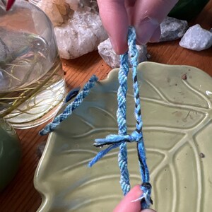 May include: A close-up of a blue and black friendship bracelet being tied. The bracelet is made of woven string and is being tied with a knot.
