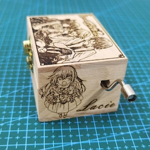 Pu&ograve; includere: Una scatola musicale in legno con una manovella sul lato. La scatola &egrave; decorata con un'immagine in bianco e nero di personaggi anime e il nome "Lacie" &egrave; inciso sul davanti.