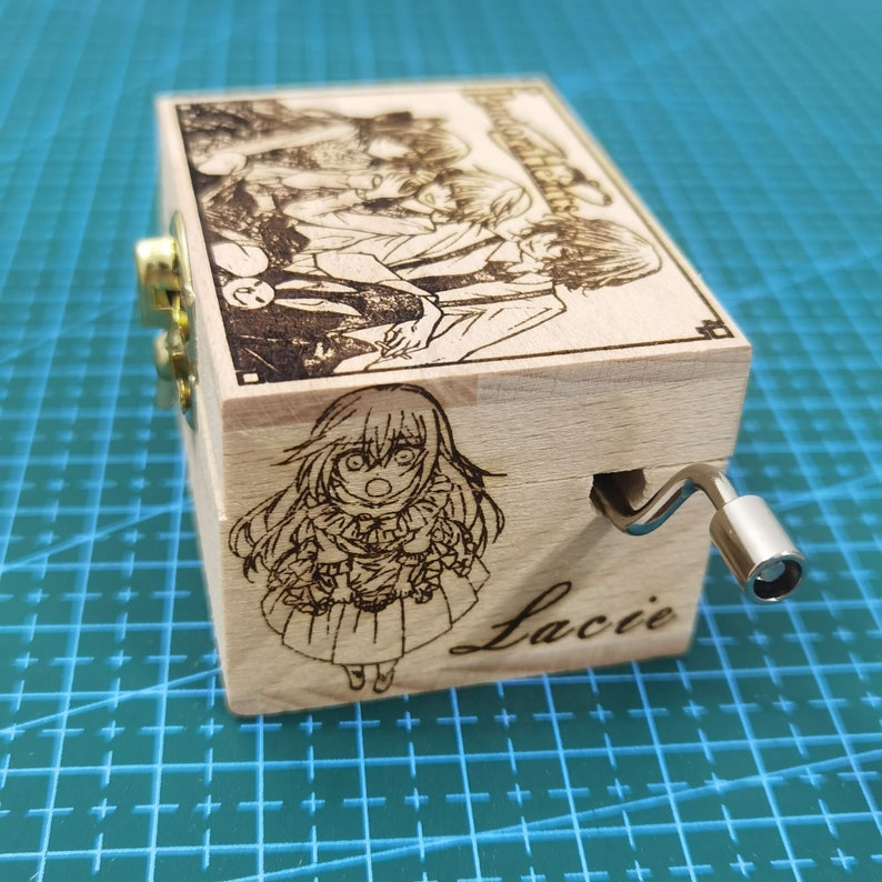 Pu&ograve; includere: Una scatola musicale in legno con una manovella sul lato. La scatola &egrave; decorata con un'immagine in bianco e nero di personaggi anime e il nome "Lacie" &egrave; inciso sul davanti.