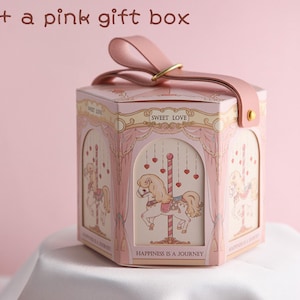 May include: A pink hexagonal gift box with a carousel design. The box has a pink ribbon and a gold metal ring. The box has the text "Sweet Love" and "Happiness is a Journey" printed on it.