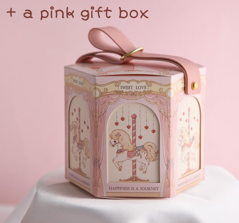 May include: A pink hexagonal gift box with a carousel design. The box has a pink ribbon and a gold metal ring. The box has the text "Sweet Love" and "Happiness is a Journey" printed on it.