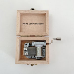 Puede incluir: Una caja de m&uacute;sica de madera con una tapa con bisagras. La tapa est&aacute; abierta y el interior de la caja es visible. La caja de m&uacute;sica tiene una manivela en el lateral y un mecanismo de m&uacute;sica de metal en el interior. La tapa tiene el texto "Here your message" grabado.