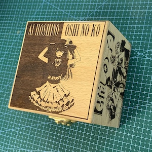 Może przedstawiać: Drewniana skrzynka z pokrywą na zawiasach, przedstawiająca laserowo grawerowany obraz postaci z anime z tekstem "Ai Hoshino Oshi no Ko".