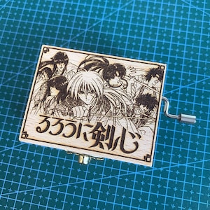 Op de afbeelding: Een houten muziekdoos met een zwart-witte illustratie van anime-personages. De muziekdoos heeft een handwindmechanisme aan de zijkant en de tekst "うっすら剣心" is op de voorkant gedrukt.