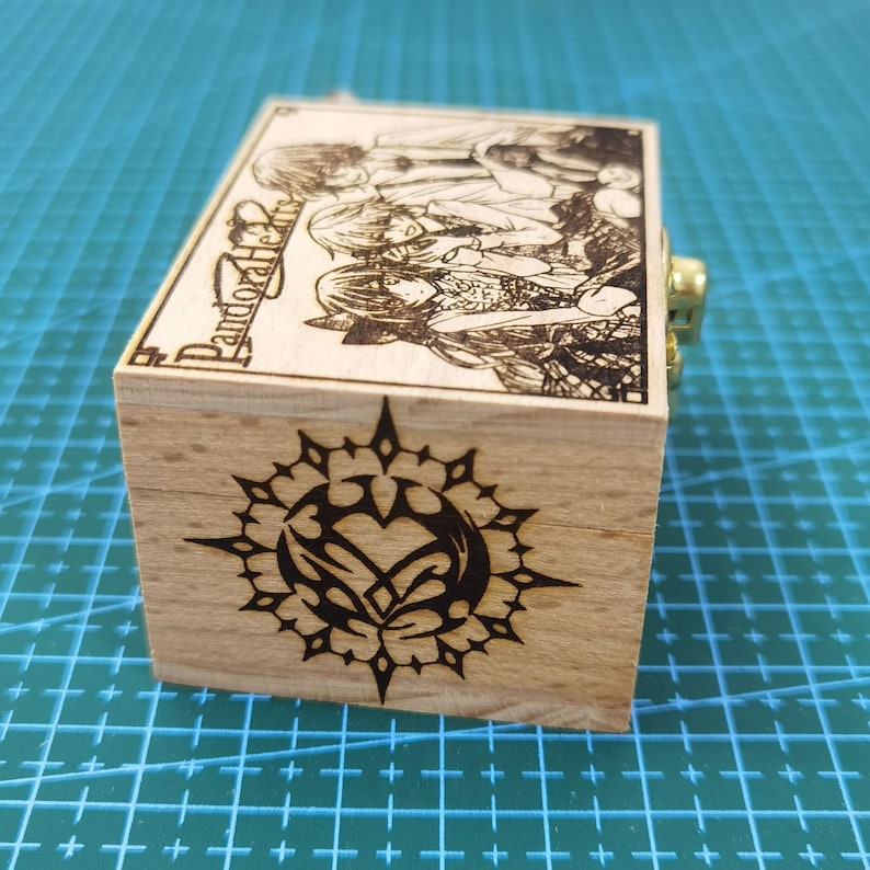 Pu&ograve; includere: Scatola musicale in legno con un design in bianco e nero di un cuore con ali all'interno di un cerchio con punte. La scatola ha una chiusura dorata e il testo "Pandora Hearts" &egrave; inciso sul lato.