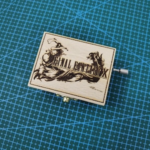 Puede incluir: Una caja de música de madera con un diseño grabado con láser que presenta un personaje de Final Fantasy VI, el texto "FINAL FANTASY VI" y las palabras "for..." en la parte inferior.