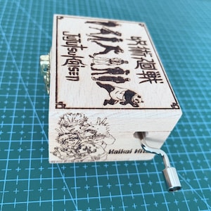 Puede incluir: Una caja de m&uacute;sica de madera con una manivela en el lateral. La caja est&aacute; decorada con una imagen en blanco y negro de personajes del anime "Jujutsu Kaisen" y el texto "Jujutsu Kaisen" y "呪術廻戦".