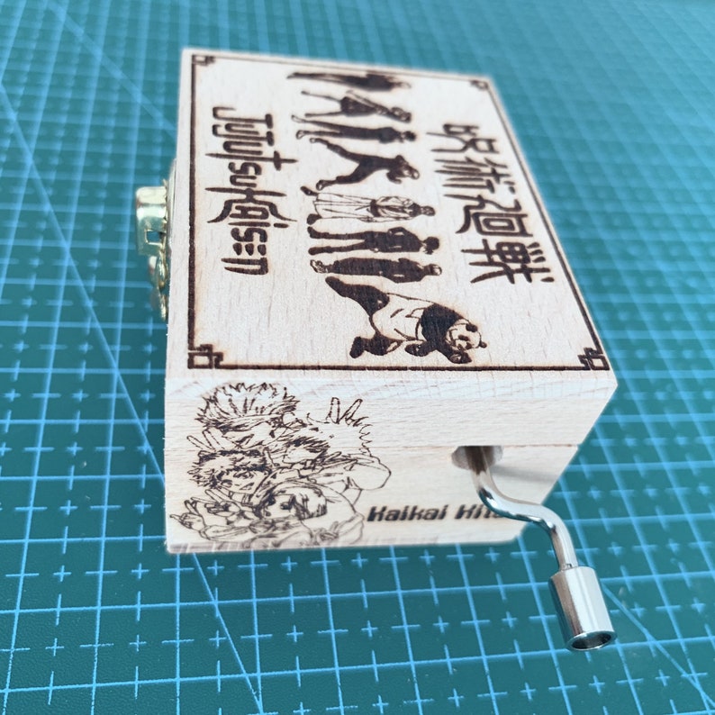 Puede incluir: Una caja de m&uacute;sica de madera con una manivela en el lateral. La caja est&aacute; decorada con una imagen en blanco y negro de personajes del anime "Jujutsu Kaisen" y el texto "Jujutsu Kaisen" y "呪術廻戦".