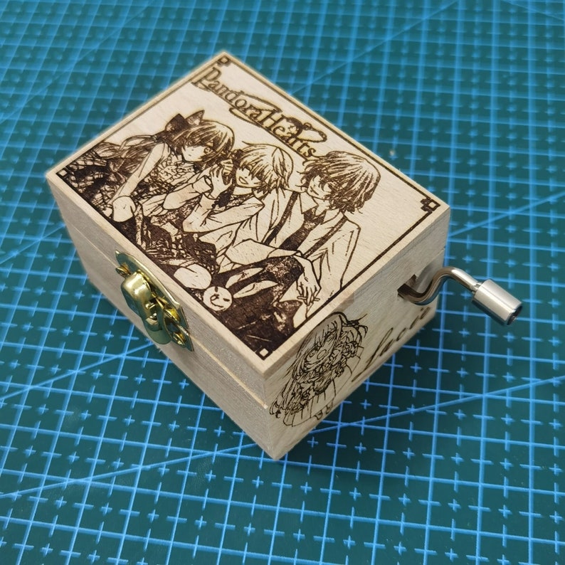 Pu&ograve; includere: Una scatola musicale in legno con una manovella sul lato. La parte superiore della scatola presenta un'immagine in bianco e nero di quattro personaggi anime della serie "Pandora Hearts".