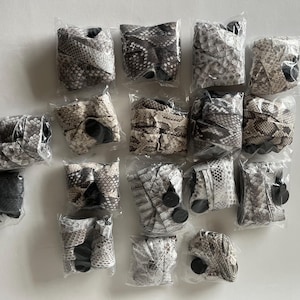 Puede incluir: Una colecci&oacute;n de mu&ntilde;equeras empaquetadas con un patr&oacute;n de piel de serpiente en varios tonos de gris, marr&oacute;n y blanco. Cada mu&ntilde;equera est&aacute; envuelta individualmente en pl&aacute;stico transparente, mostrando la textura y el dise&ntilde;o detallados.