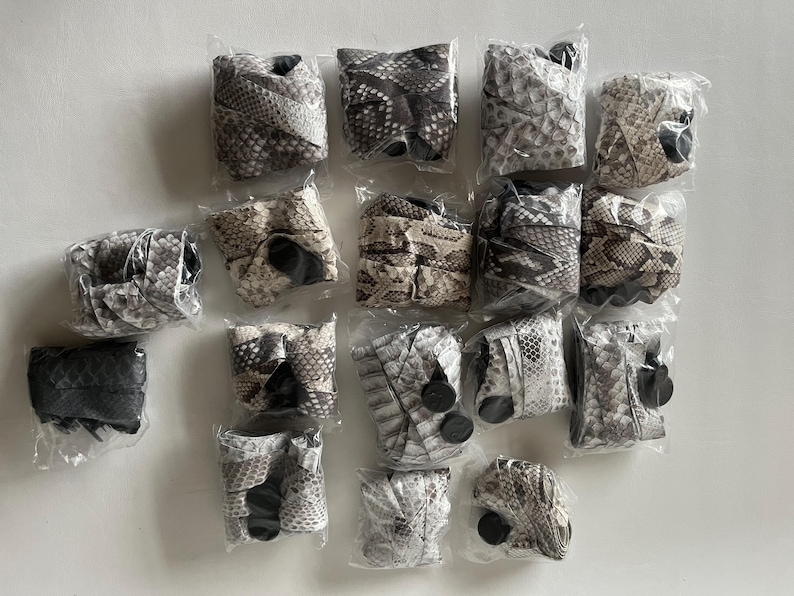 Puede incluir: Una colecci&oacute;n de mu&ntilde;equeras empaquetadas con un patr&oacute;n de piel de serpiente en varios tonos de gris, marr&oacute;n y blanco. Cada mu&ntilde;equera est&aacute; envuelta individualmente en pl&aacute;stico transparente, mostrando la textura y el dise&ntilde;o detallados.