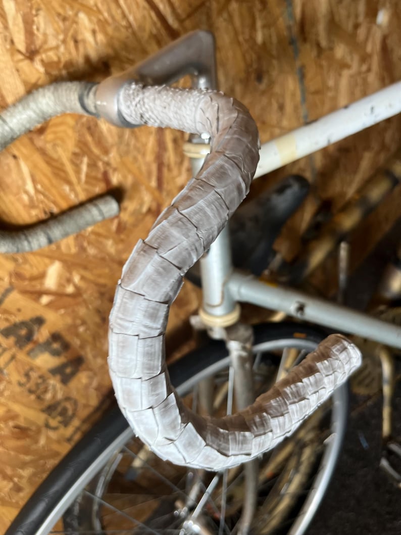 Puede incluir: Primer plano de un manillar de bicicleta con cinta texturizada de color gris claro. El manillar forma una curva en forma de U, con una parte del cuadro y la rueda de la bicicleta visibles en el fondo. La cinta parece estar despeg&aacute;ndose en algunas zonas.