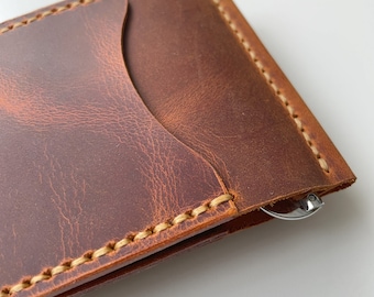 May include: A brown leather wallet with a money clip. The wallet is made of distressed leather and features a contrasting beige stitching around the edges. The wallet has a pocket on the front.