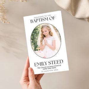 May include: A white baptism invitation with the text "WELCOME TO THE BAPTISM OF" at the top. A photo of a young girl in a white dress is in the center. The text "EMILY STEED" is below the photo, with additional details.