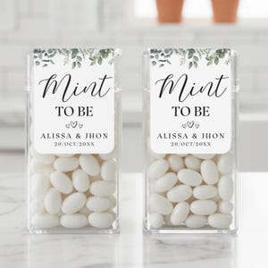 May include: Two clear plastic boxes filled with white mints. Each box has a label that reads "Mint to Be" with names and a date, and is decorated with green leaves. A wedding favor.