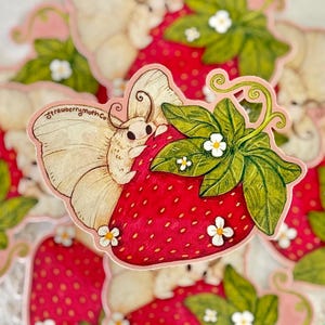 May include: A sticker featuring a whimsical illustration of a moth perched on a vibrant red strawberry. The strawberry has green leaves and small white flowers. The sticker has the text "strawberrymoth Co." in a cursive font.