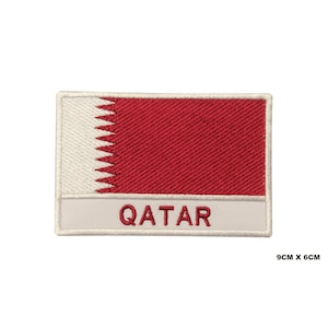 Puede incluir: Un parche bordado con la bandera de Qatar. El parche es blanco con un rectángulo granate y una línea blanca dentada. Las palabras "QATAR" están escritas en blanco debajo de la bandera.