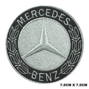 May include: A black and silver embroidered patch with the Mercedes-Benz logo. The logo is a three-pointed star inside a circle with the words "Mercedes-Benz" around the outside. The patch is 7.5 inches by 7.5 inches.