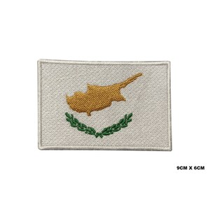 May include: White embroidered patch with the flag of Cyprus. The flag features a yellow outline of the island of Cyprus and a green olive branch wreath. The patch measures 9 inches by 6 inches.