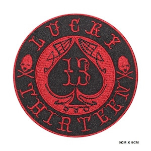 May include: Round black patch with red embroidered text and design. The design includes a red spade with the number 13, two skulls with crossbones, and the words "LUCKY THIRTEEN". The patch measures 9cm x 9cm.