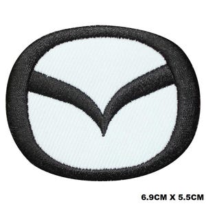 May include: Black and white embroidered patch featuring a stylised logo of a car brand. The logo is a white oval with a black, curved, wing-like design inside. The patch measures 6.9 cm by 5.5 cm.