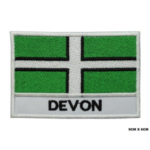May include: Embroidered patch featuring the flag of Devon, England. The rectangular patch has a green background with a white cross and black border. The word "DEVON" is embroidered in black on a white banner. The patch measures 3.5 inches by 2.4 inches.