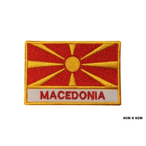 May include: Embroidered patch featuring the flag of Macedonia. The flag is red with a yellow sun in the center and eight yellow rays extending from the sun. The patch is bordered in yellow and has the word "MACEDONIA" written in yellow on a white background.
