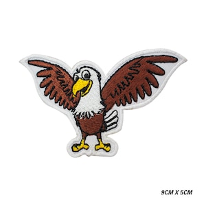 May include: An embroidered patch featuring a cartoon eagle with brown wings, a white head, and a yellow beak and feet. The eagle is depicted with its wings spread wide. The patch measures 9cm x 5cm as indicated by the text in the corner.