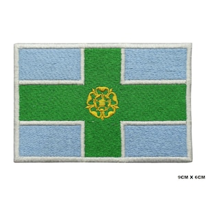 May include: An embroidered patch featuring the flag of Greater Manchester, England. The flag is a white cross on a blue background with a green cross over the top. The green cross has a gold rose in the centre. The patch is 9 cm by 6 cm.