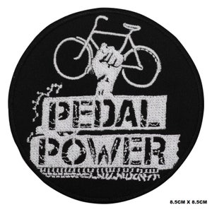 Puede incluir: Parche bordado en blanco y negro con una bicicleta y un puño en alto. El texto "PEDAL POWER" está debajo de la bicicleta. El parche mide 8,5 cm por 8,5 cm.
