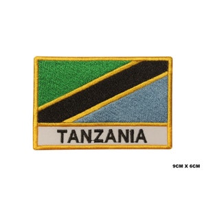 May include: Embroidered patch depicting the Tanzanian flag. The rectangular patch features a green upper section, a black diagonal stripe, and a light blue lower section. The word "TANZANIA" is embroidered in black on a white banner. Measures 9cm x 6cm.