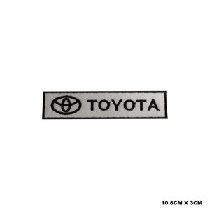 Peut inclure: Écusson brodé blanc avec inscription noire "TOYOTA" et un logo stylisé noir d'un cercle avec une ovale à l'intérieur.