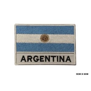 May include: An embroidered patch featuring the Argentinian flag with the words "ARGENTINA" below. The flag is blue and white with a yellow sun in the center. The patch is 9 cm by 6 cm.
