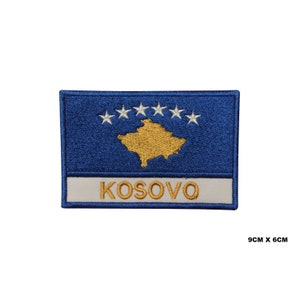 May include: Embroidered patch featuring the flag of Kosovo. The patch is blue with a gold outline and six white stars in a circle. The word "KOSOVO" is written in gold below the flag.