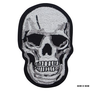 Puede incluir: Parche bordado blanco en forma de calavera sobre fondo negro. La calavera tiene un diseño detallado con la frente agrietada y una sonrisa de dientes. El parche mide 6 cm por 9 cm.