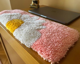 May include: A pink and white fuzzy wrist rest with a yellow daisy design. The wrist rest is on a wooden surface.