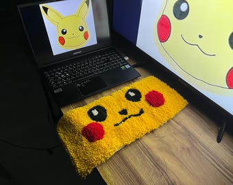 Può includere: Un tappeto di peluche giallo e rosso con un viso di cartone animato di Pikachu, un personaggio popolare di Pokemon. Il tappeto è su una superficie di legno.