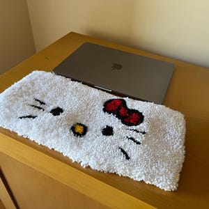 Puede incluir: Alfombrilla blanca y esponjosa para portátil con un diseño de Hello Kitty con ojos negros, un lazo rojo y una nariz amarilla.