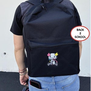 May include: Black backpack with a zippered pocket and a small embroidered design of a white robot with pink and yellow accents. The text "Back 2 School" is written in a red circle on the backpack.